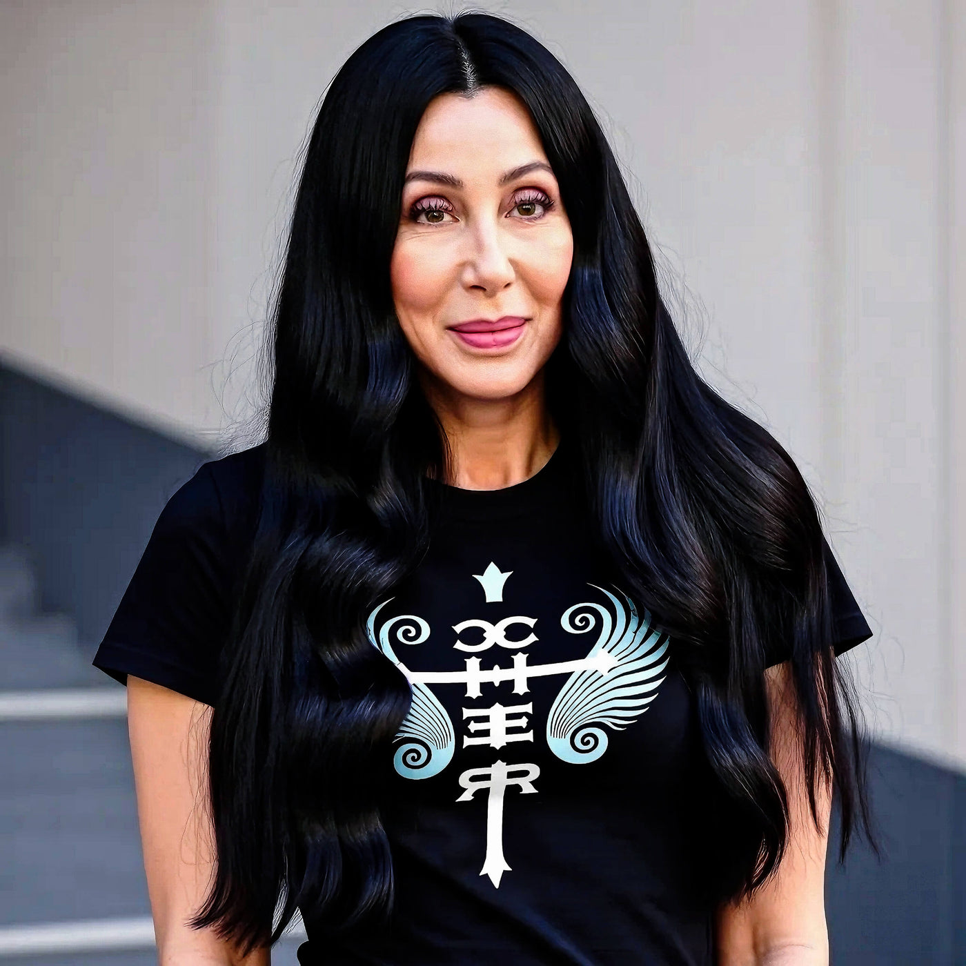 Cher with long, wavy black hair, wearing a black Cher Fan Club T-shirt designed by Gllamazon, featuring a stylized design with her name and wing-like flourishes.