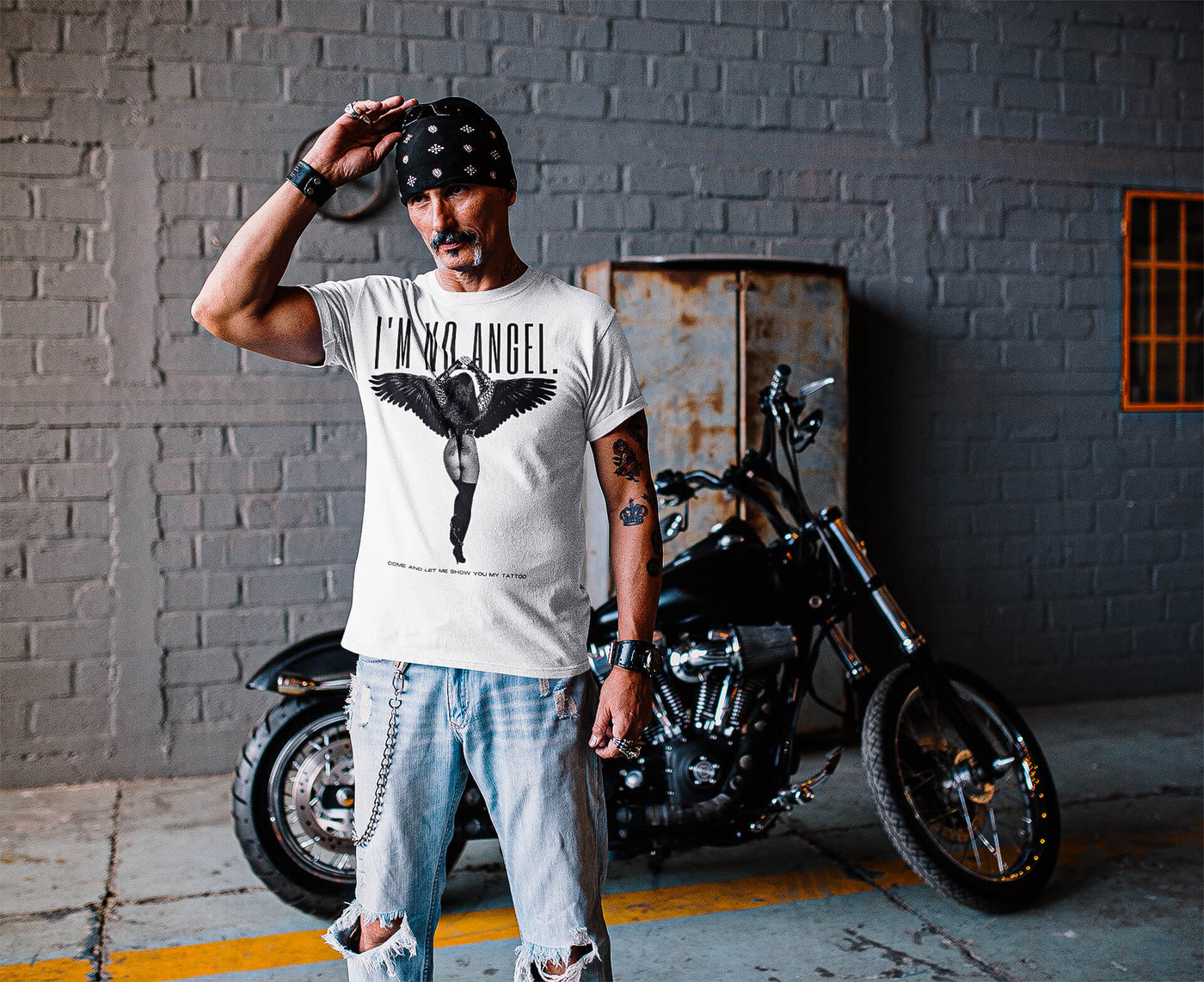 Biker with a bandana, stylishly wearing 'I'm No Angel Cher' T-shirt by Gllamazon, posing confidently in front of his motorcycle.