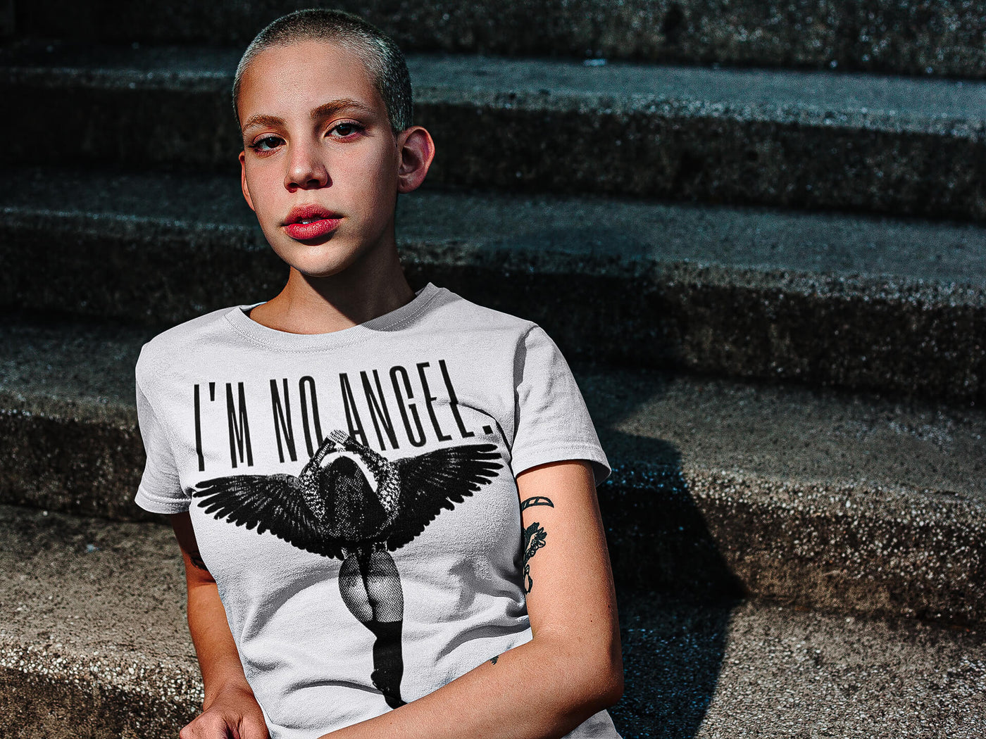Beautiful young woman with a buzz cut, casually sitting on street stairs, wearing 'I'm No Angel Cher' T-shirt by Gllamazon, embodying urban style.