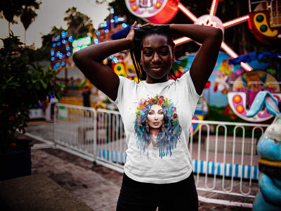 Black young woman with dreadlocks near a carousel wearing Gllamazon's Cher-ry Blossom Cher T-shirt. Color: White.
