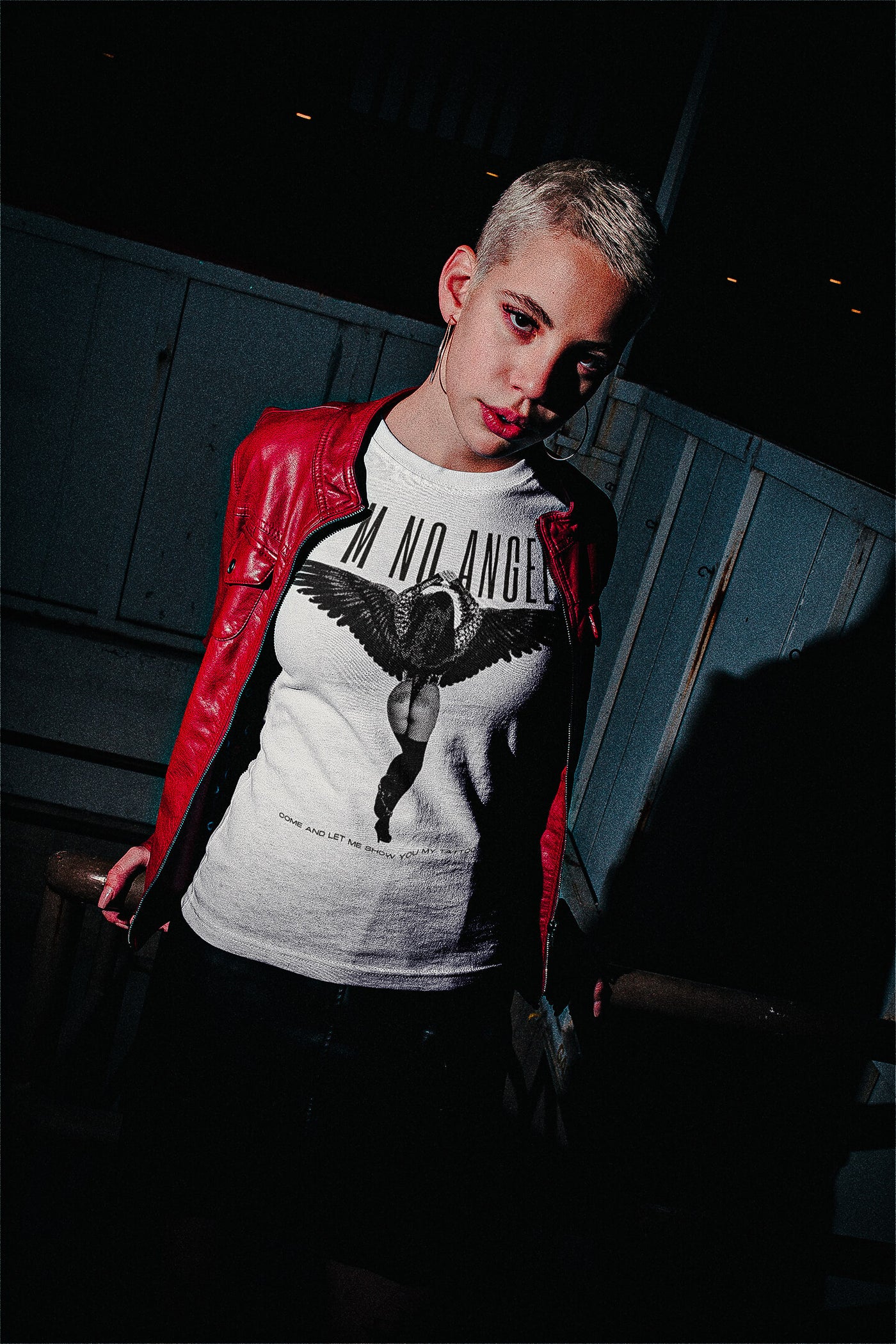 Short-haired woman in a night setting, sporting 'I'm No Angel Cher' T-shirt by Gllamazon in a mockup style, blending fashion with urban nightlife.