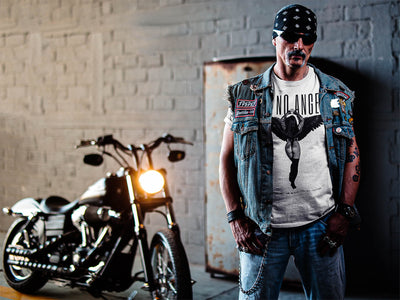 Tattooed biker man with bandana and chain, wearing 'I'm No Angel Cher' T-shirt by Gllamazon, in front of a motorcycle, exuding rugged charm.
