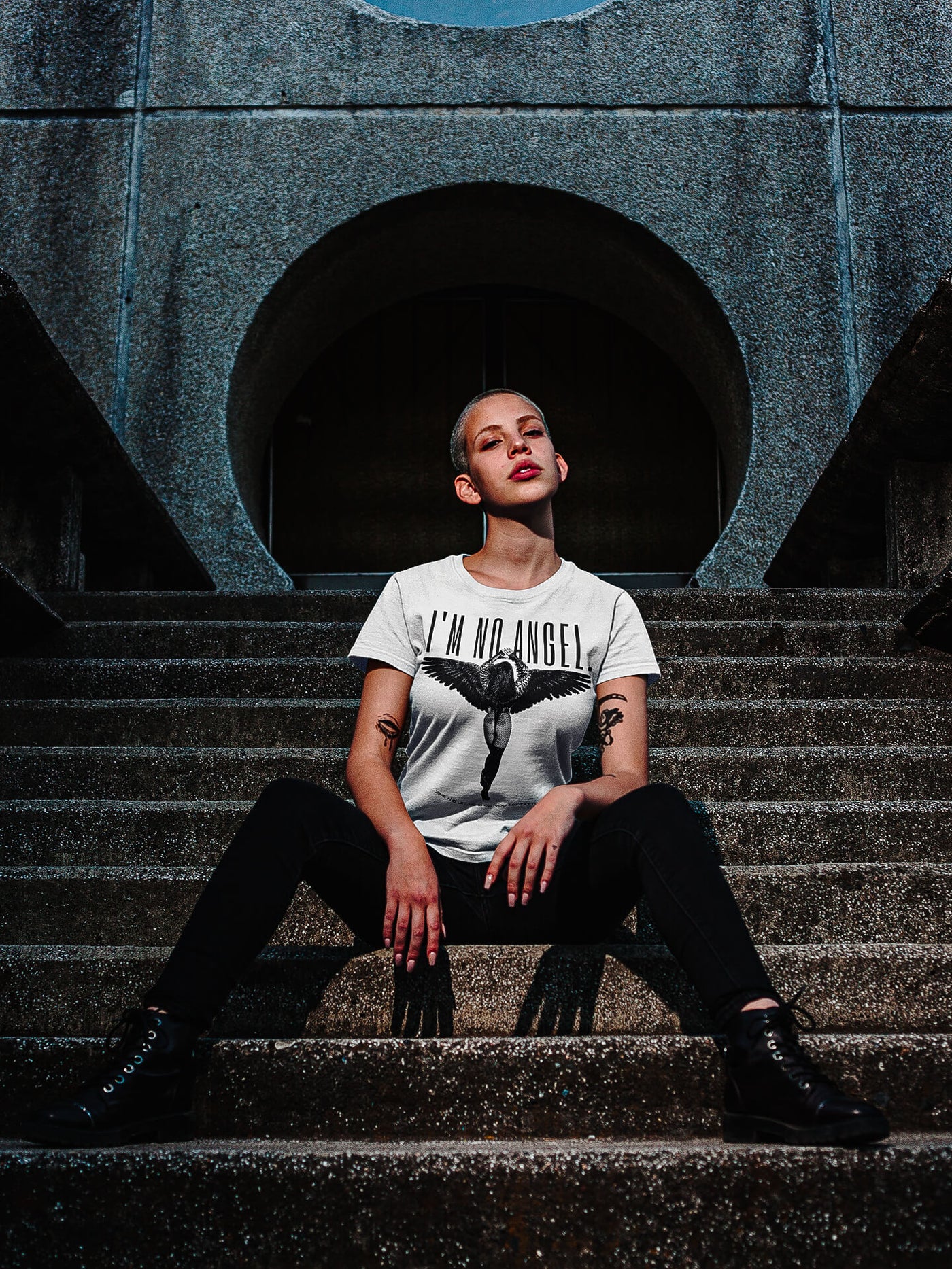 Trendy girl with tattoos, sitting on street stairs, wearing 'I'm No Angel Cher' T-shirt by Gllamazon, showcasing a blend of street fashion and individuality.